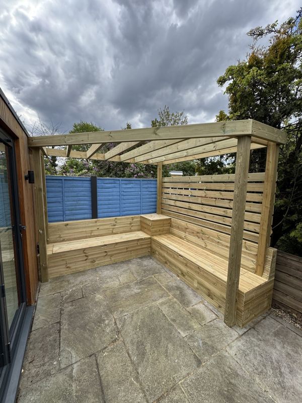 L-shaped decking benches with storage built into the corner of a patio, under a pergola frame